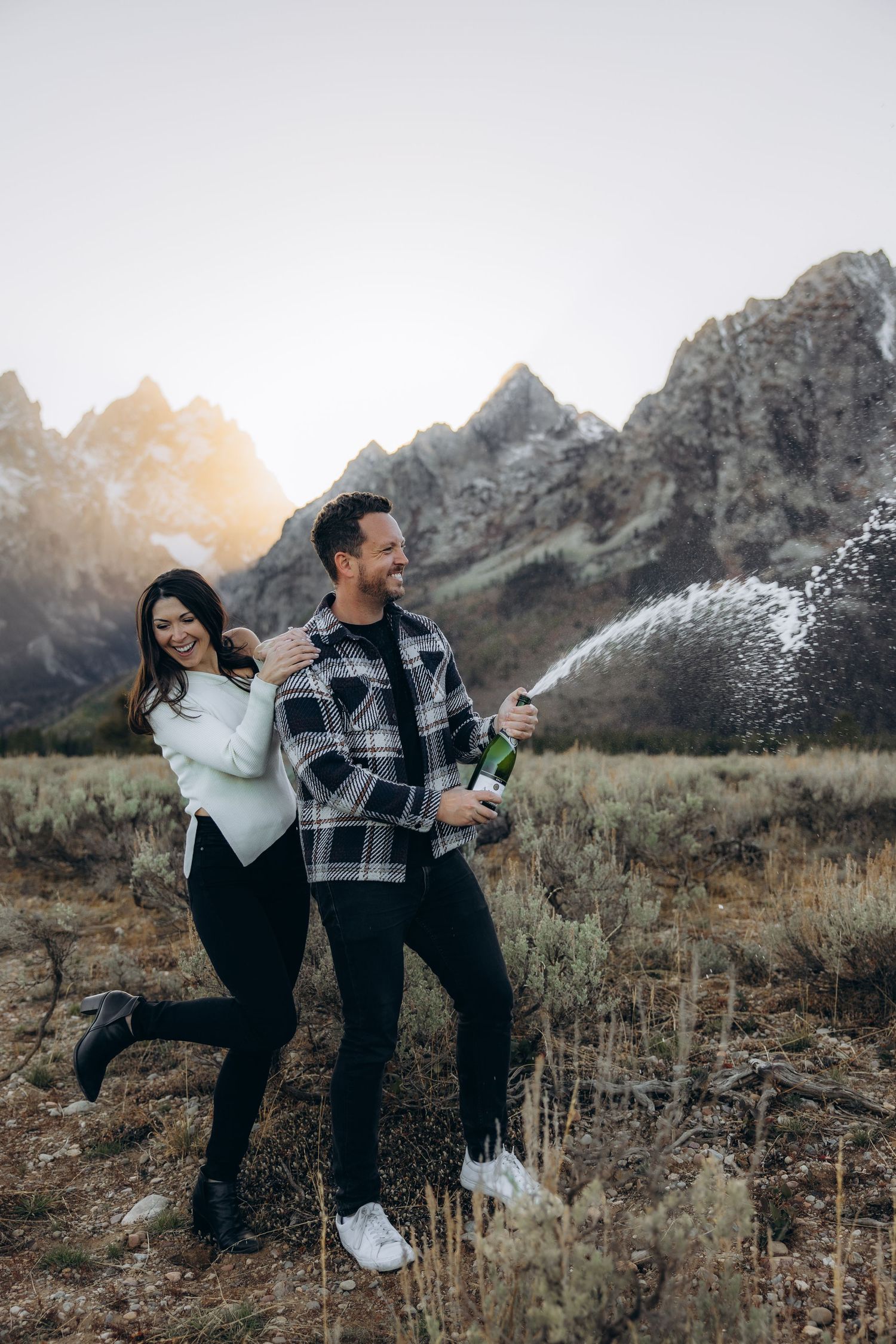 A couple sprays champagne while celebrating among the Grand Teton mountains at sunset.