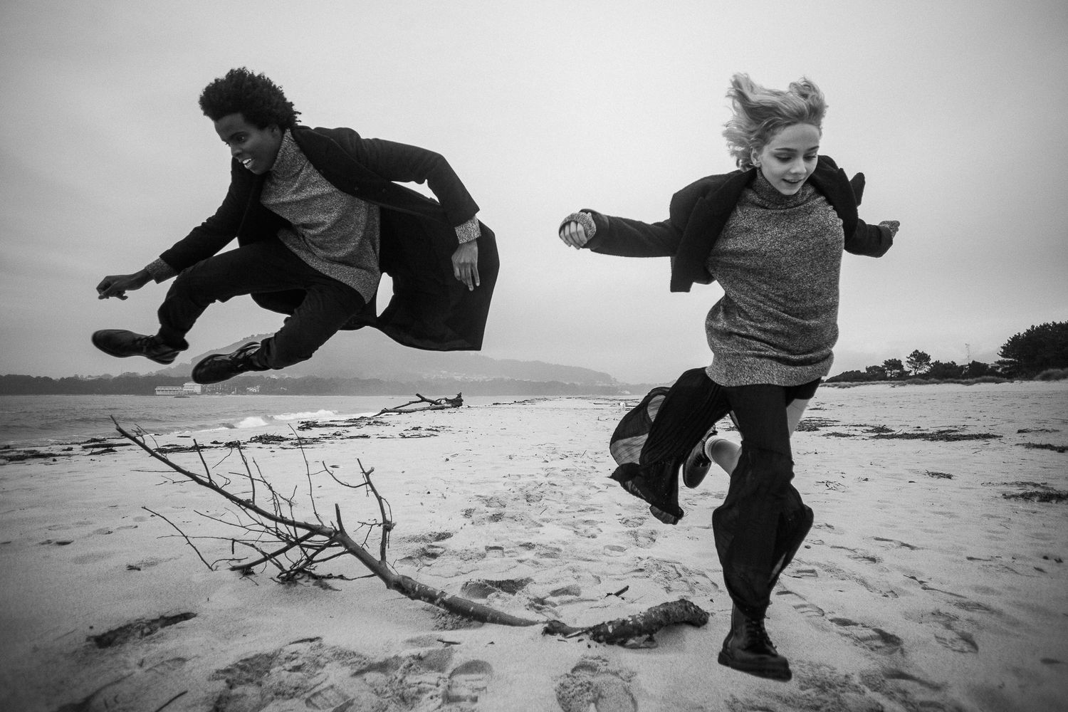 Two people jump together on a windswept beach in dramatic black and white photography.