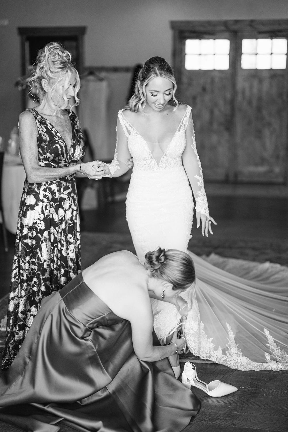 Black and white wedding photo showing final dress preparations with attendants helping adjust the bride's long lace train.