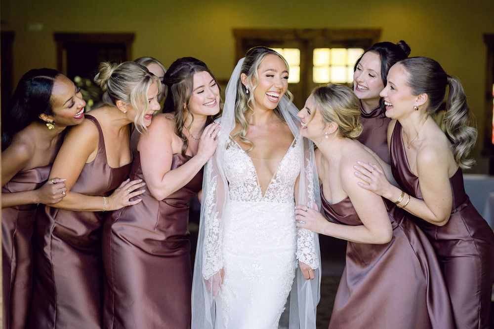 Bridesmaids in dusty rose dresses share a joyful moment with bride wearing white lace gown during wedding preparations.