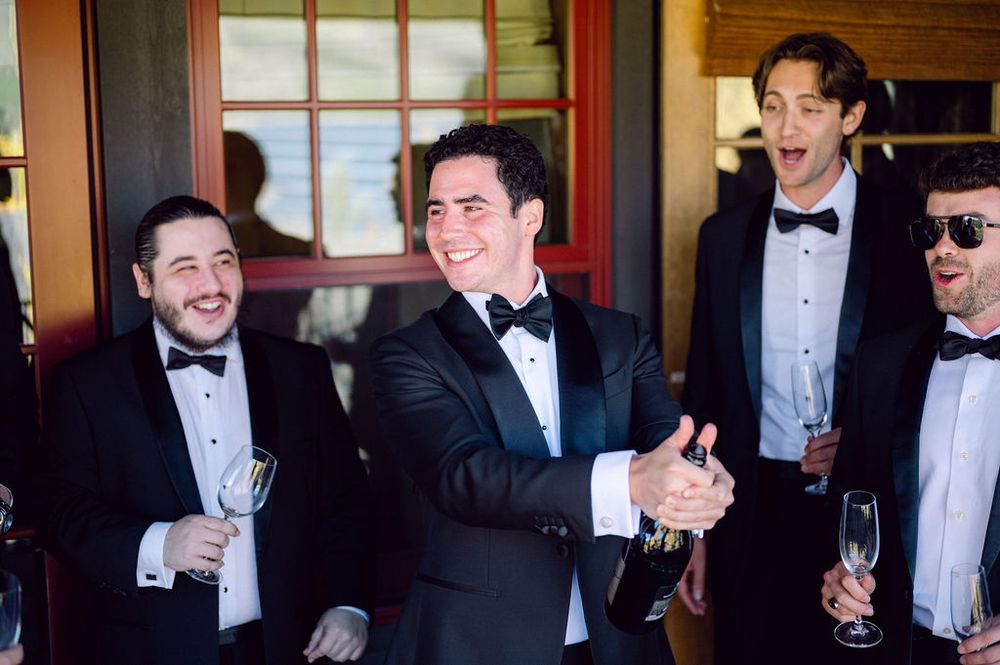 A group of men in formal black tuxedos and bow ties share laughs and drinks at an elegant event.