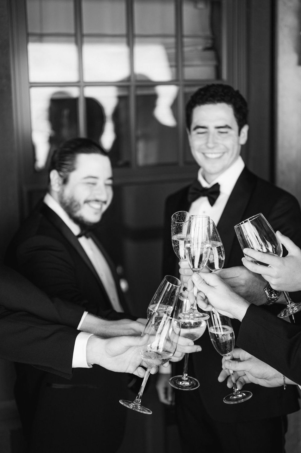Groomsmen in black suits raise champagne glasses for a celebratory toast in black and white photography.