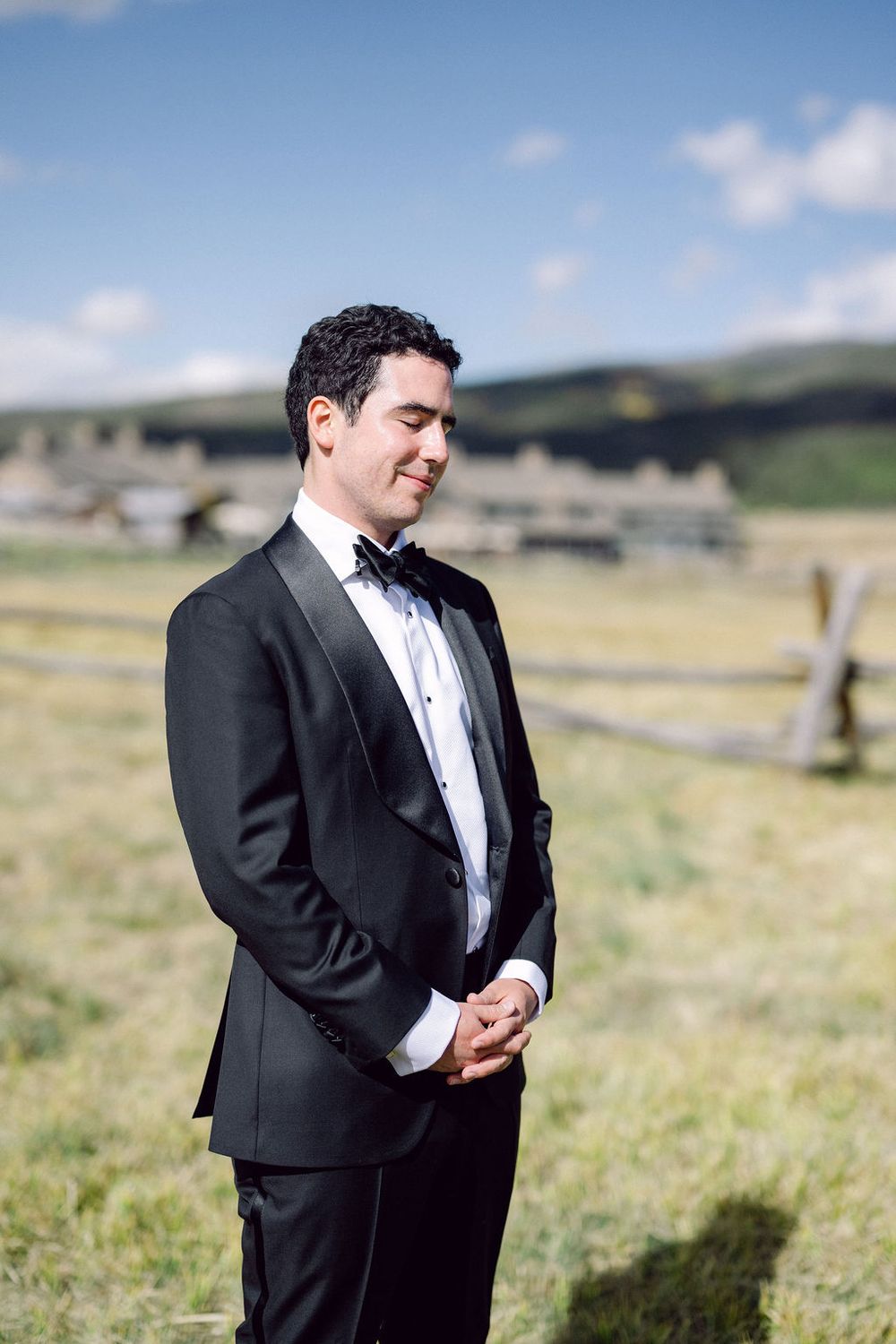 A groom in a classic black tuxedo and bowtie stands in a sunny mountain meadow with rolling hills in the background.