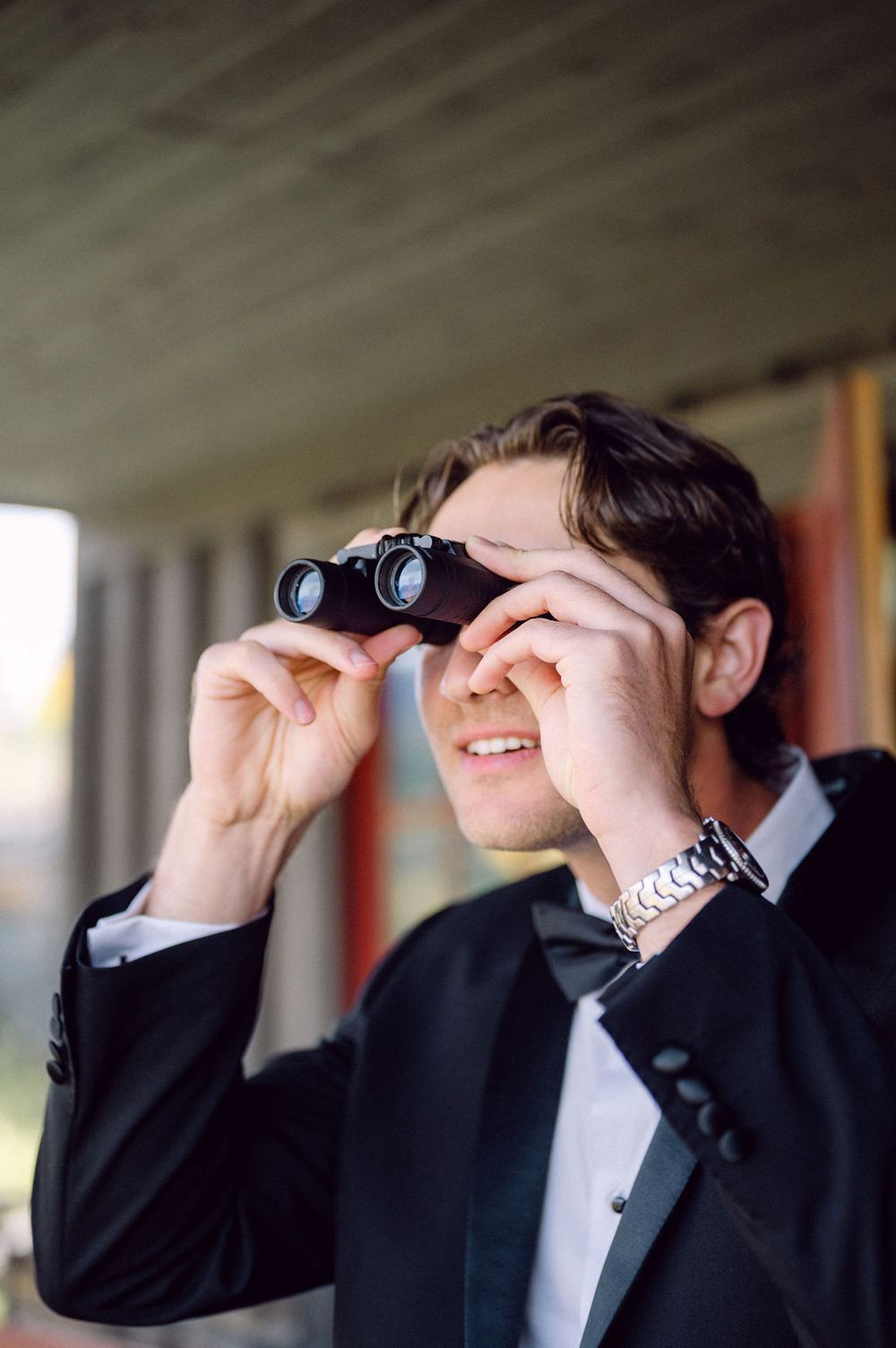 A groomsman in a black suit and tie looks through binoculars in a sequence of candid wedding party photos.