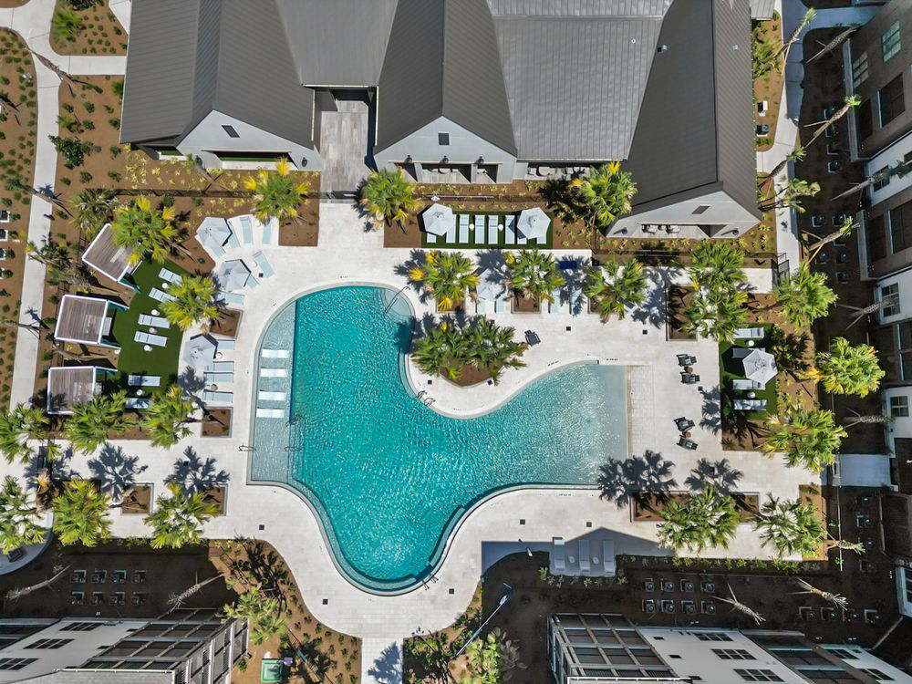 Aerial view of a luxury resort swimming pool surrounded by palm trees and lounge chairs between modern buildings.