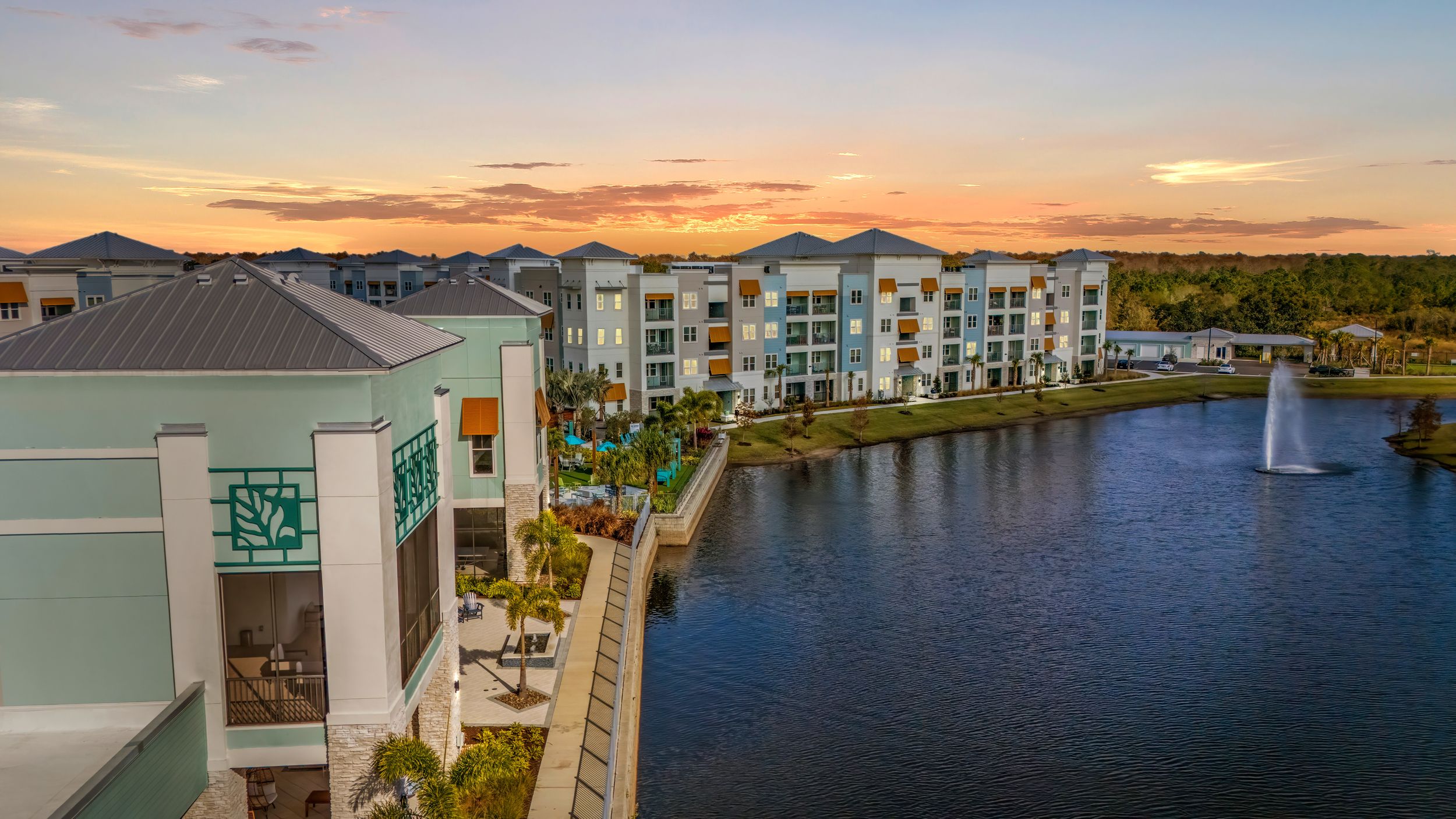 Waterfront condominiums with pastel colors reflect in calm waters during a vibrant sunset over coastal mountains.