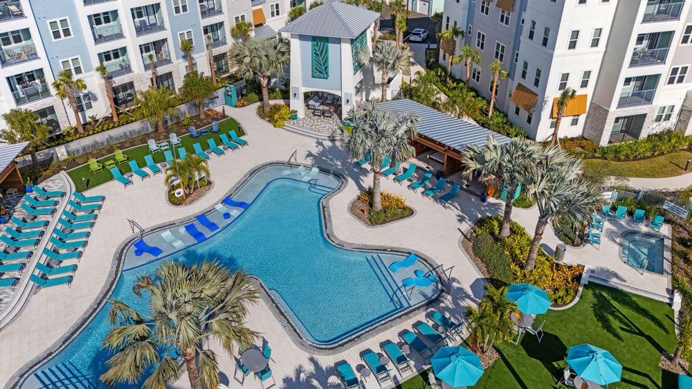 Aerial view of a luxury resort pool area with turquoise loungers and white gazebo surrounded by apartment buildings.