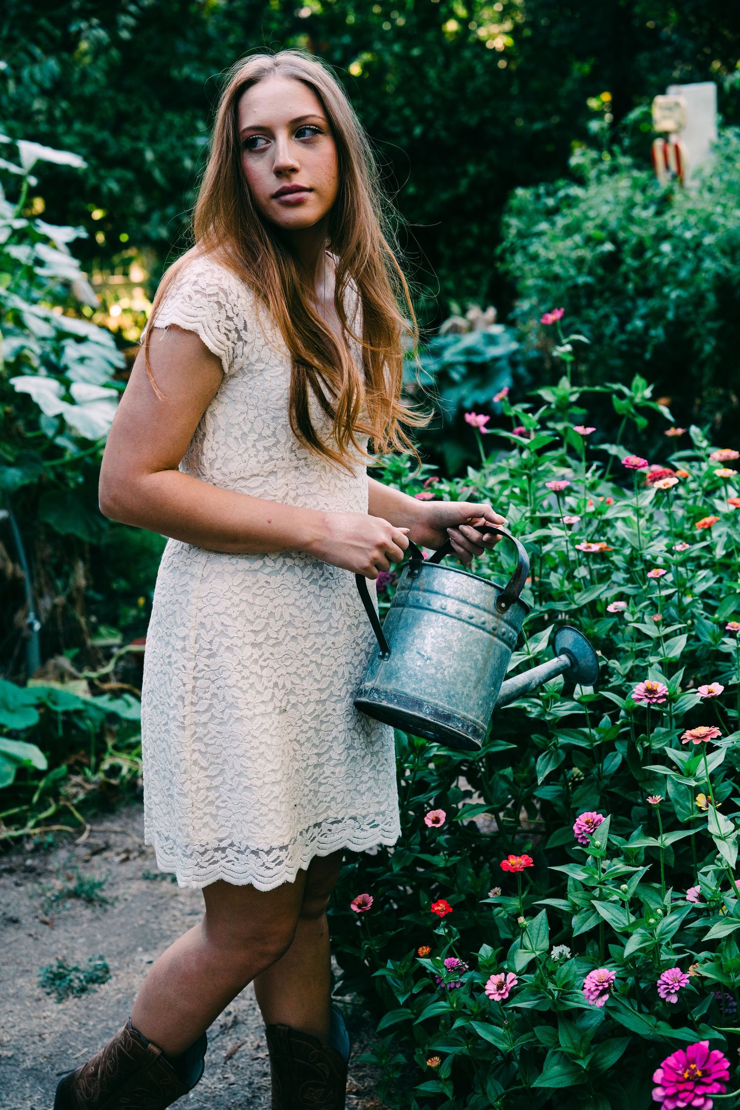 A young woman in a lace dress holds a watering can in a flourishing garden.