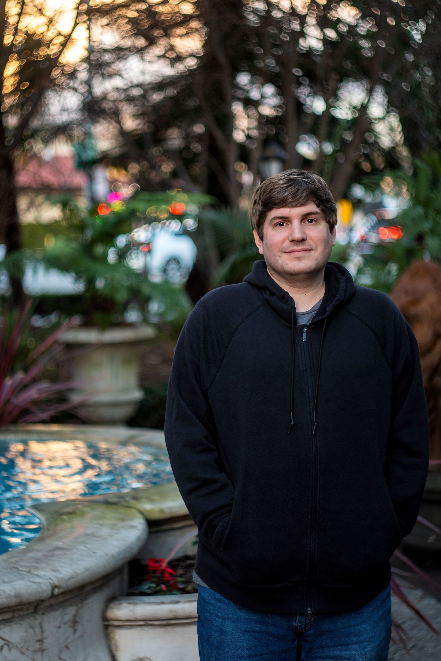 Person in black hoodie standing by ornate fountain at dusk.
