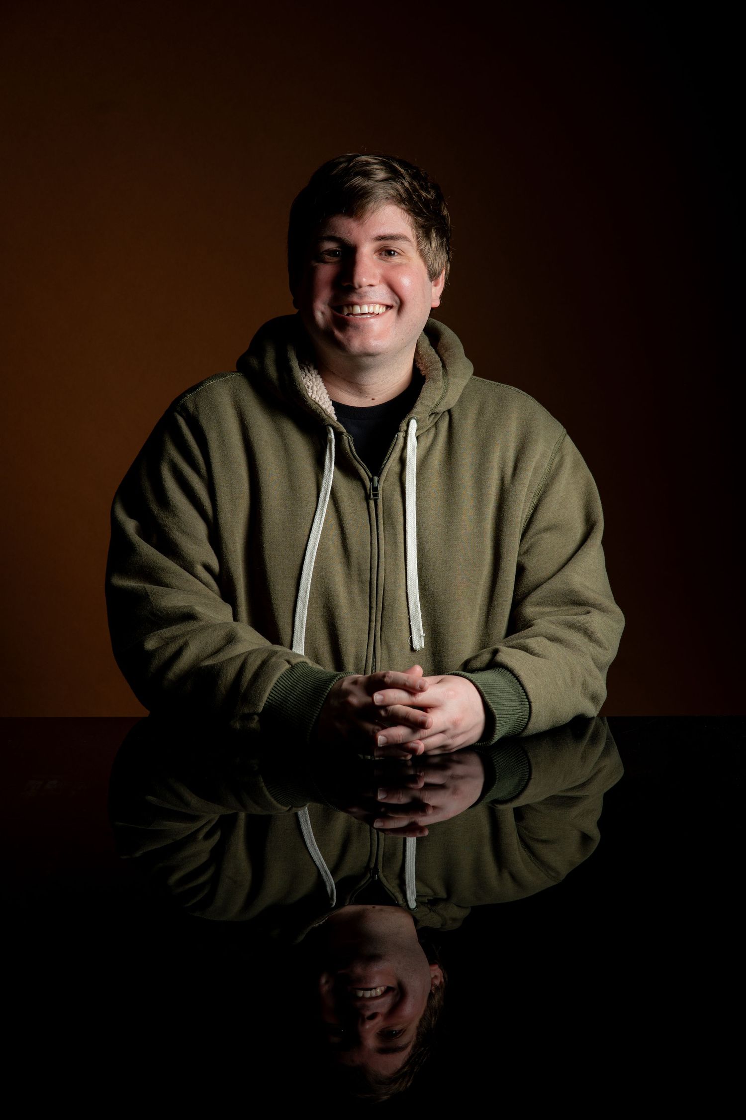 Person in olive green hoodie sitting at reflective table in moody lighting.