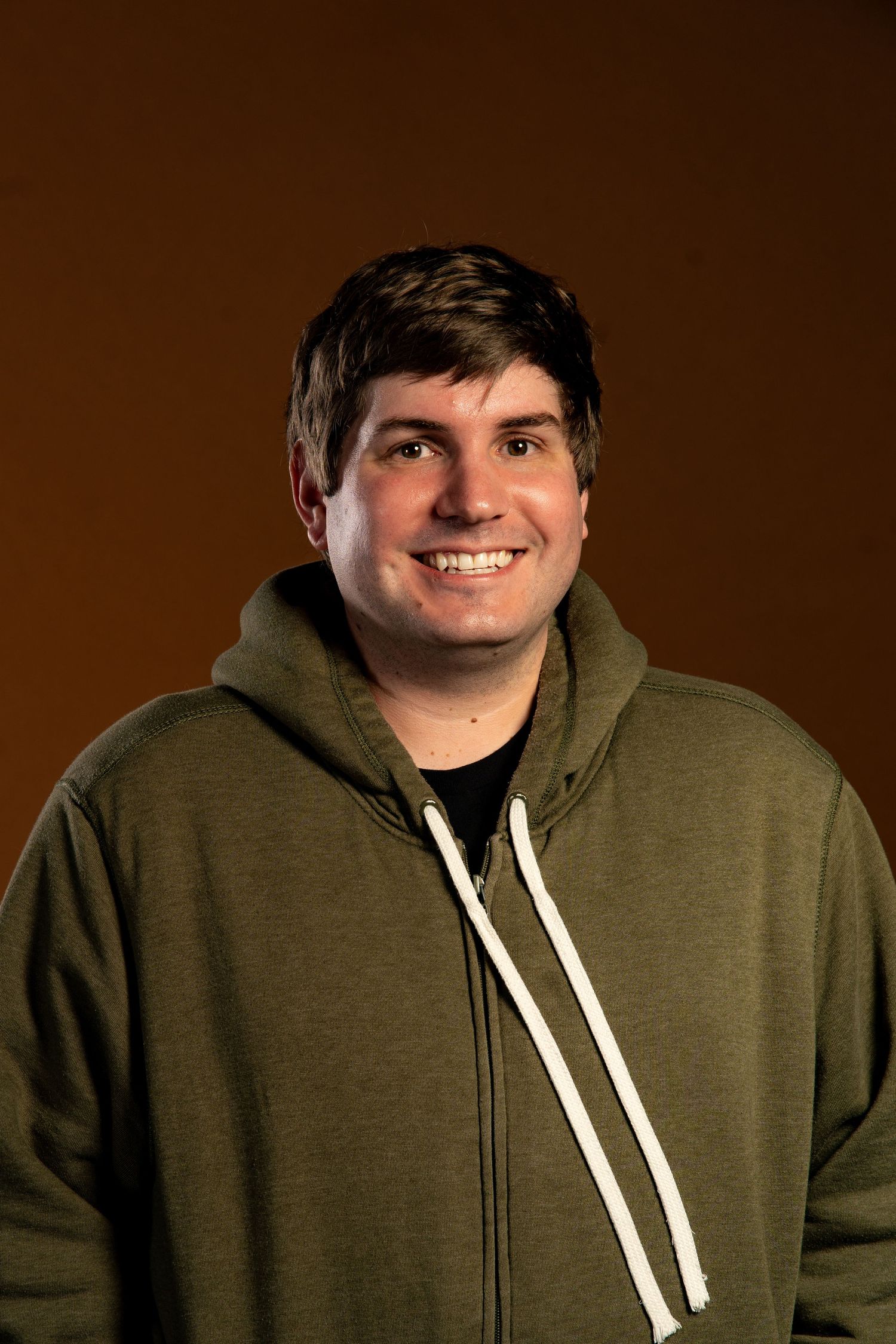 Portrait of someone wearing an olive green zip-up hoodie against a brown background.