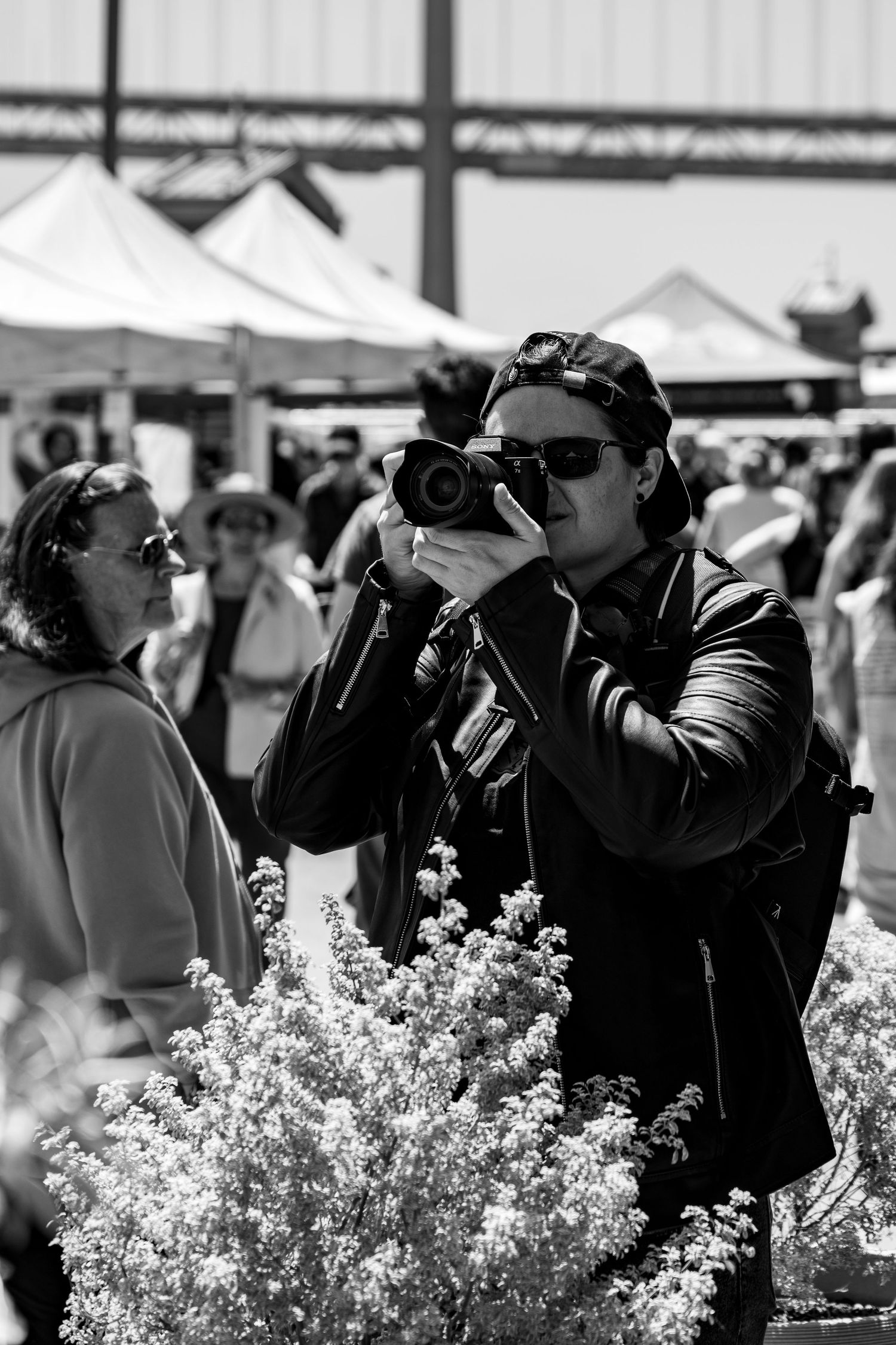 Photographer in dark clothing taking pictures at an outdoor market with white tents and flowers.