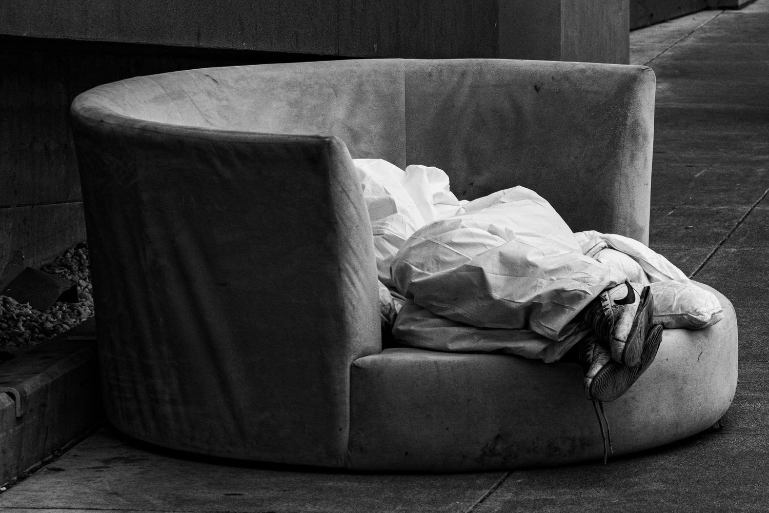 A black and white photo of a homeless man sleeping on a curved chair with crumpled fabric.