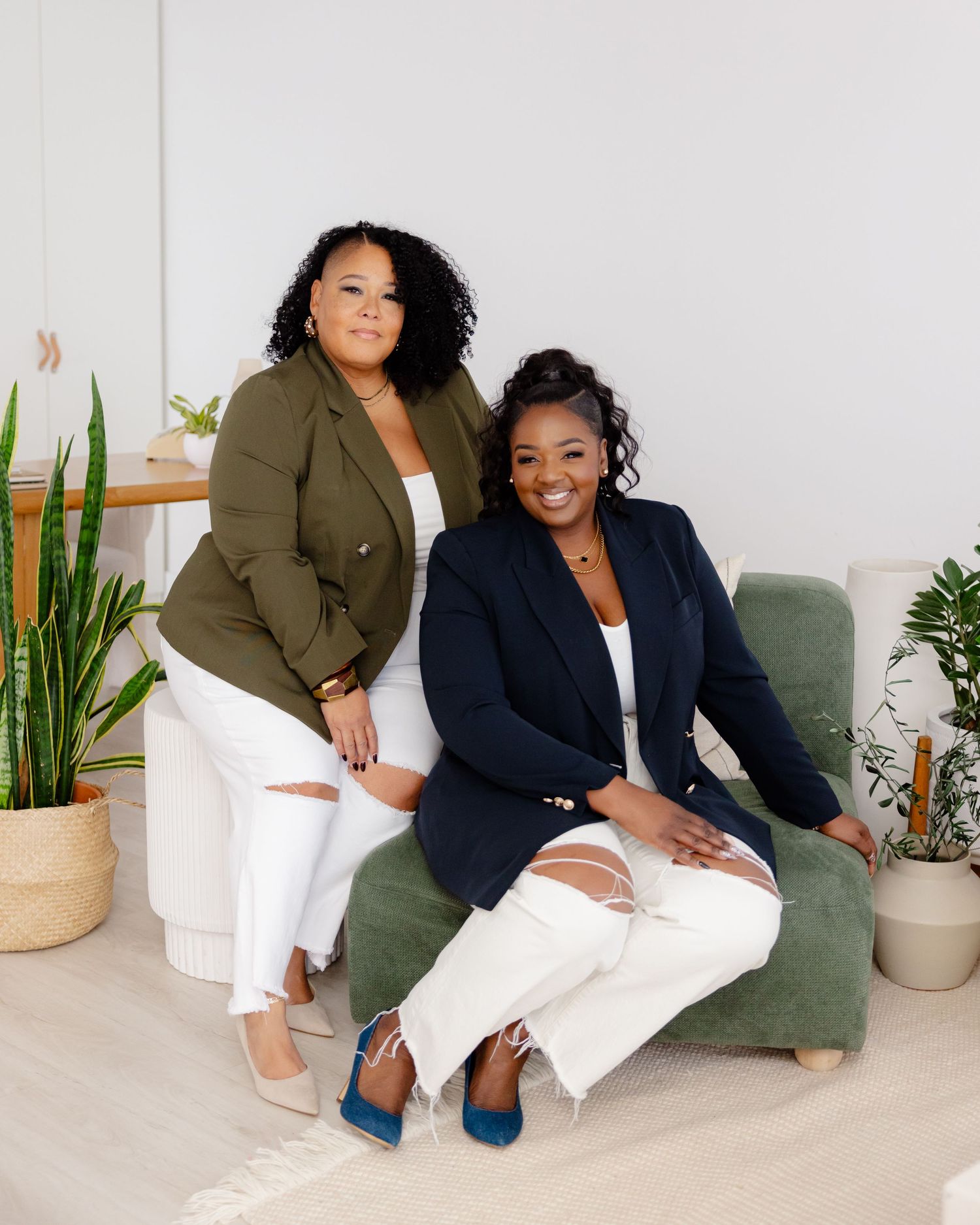 Two women in business attire sit together on a green couch surrounded by potted plants in a bright studio setting.