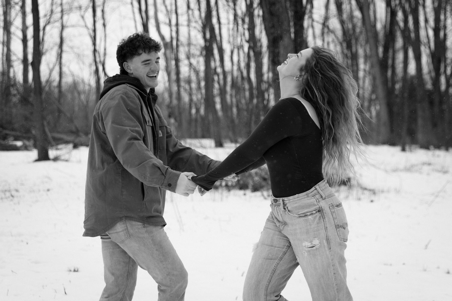 A playful couple dance and spin together in a snowy winter forest setting captured in black and white.