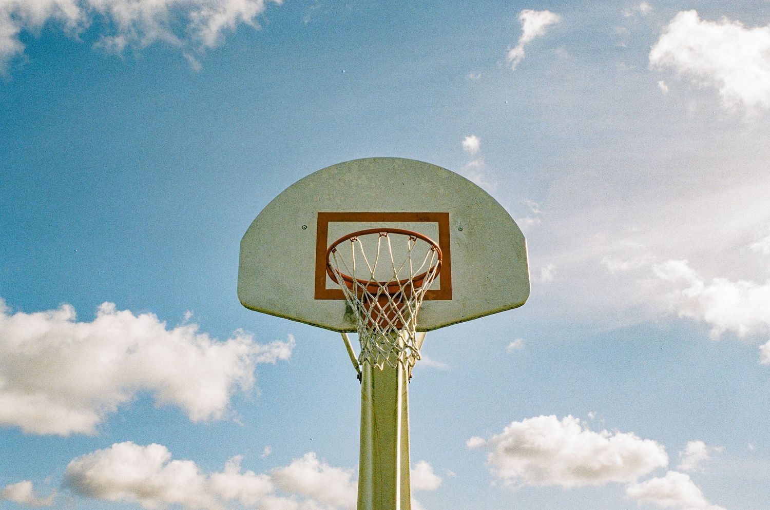 Basketball hoop against blue sky with white fluffy clouds in natural lighting.