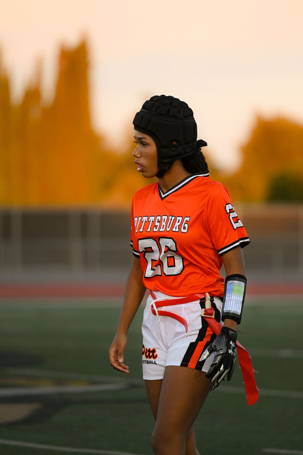 Player in orange and white uniform stands on sports field at sunset.