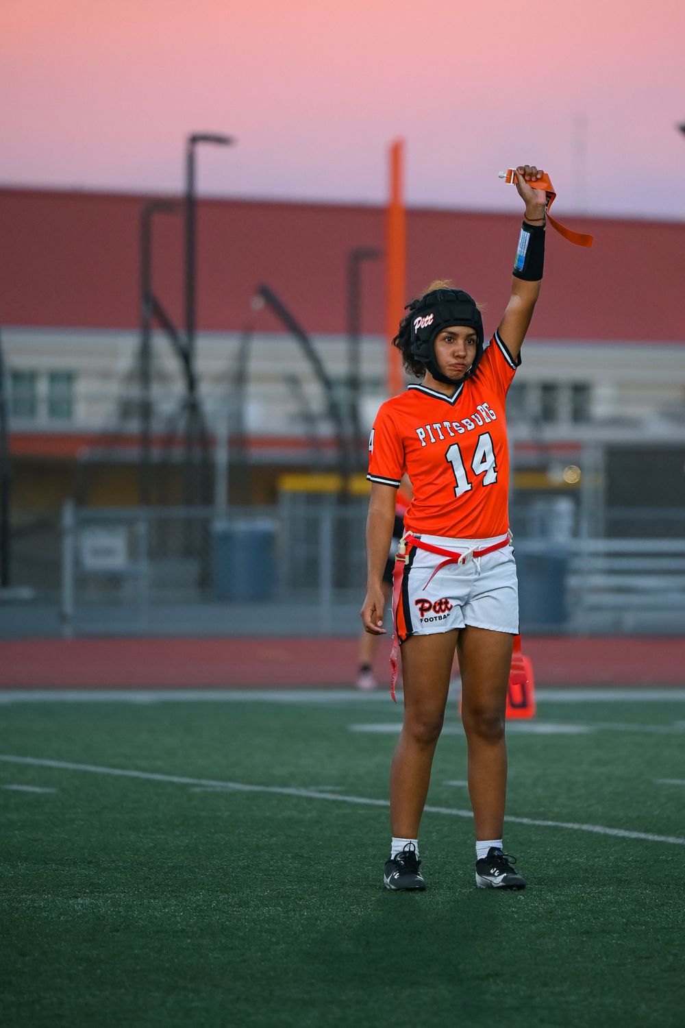 Player in orange jersey number 14 stands ready on football field at dusk.