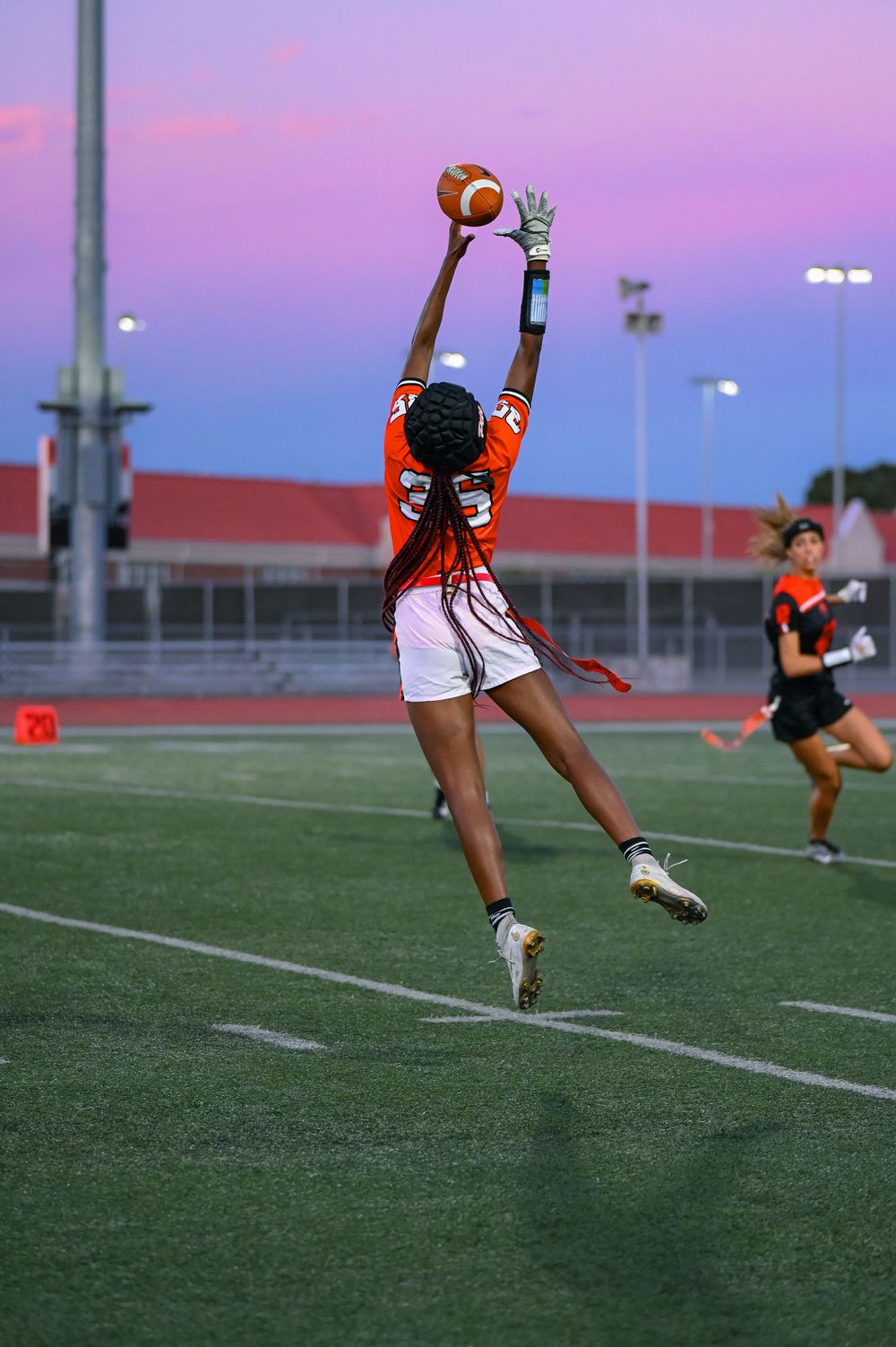 Player in orange jersey leaps high to catch football against purple sunset sky.