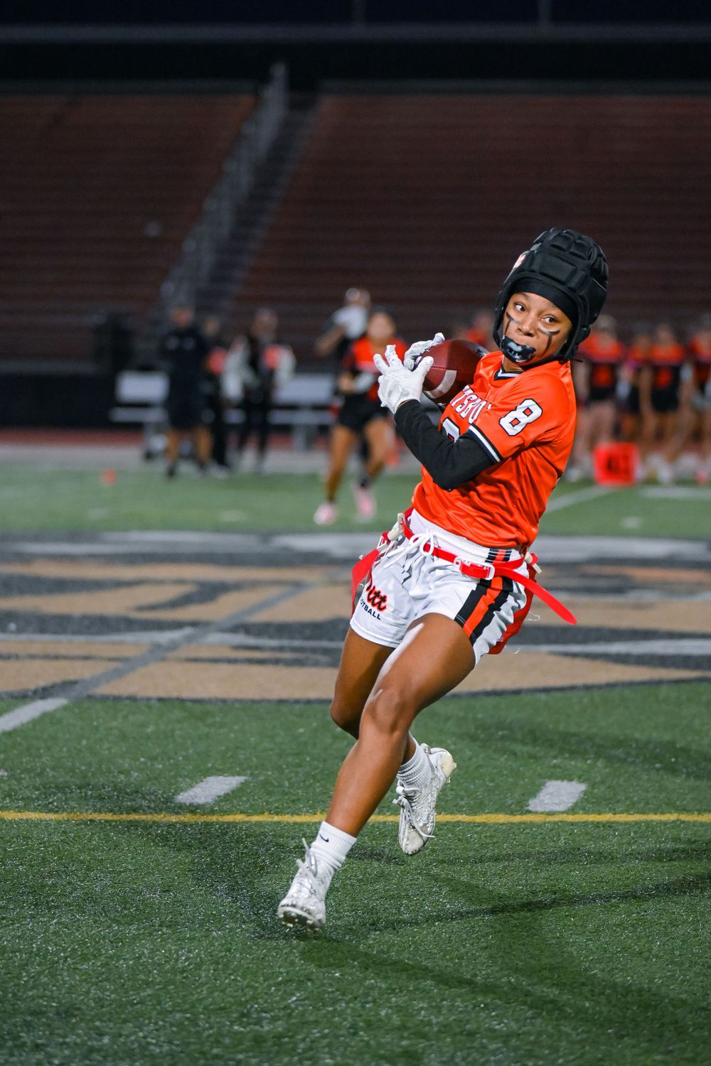 Player in orange uniform catches football during evening practice.