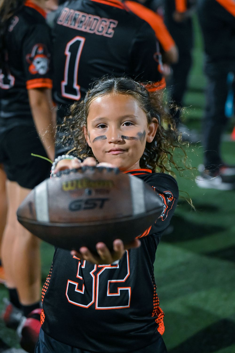 Youth football player in black and orange uniform holds up a GST football during a game.