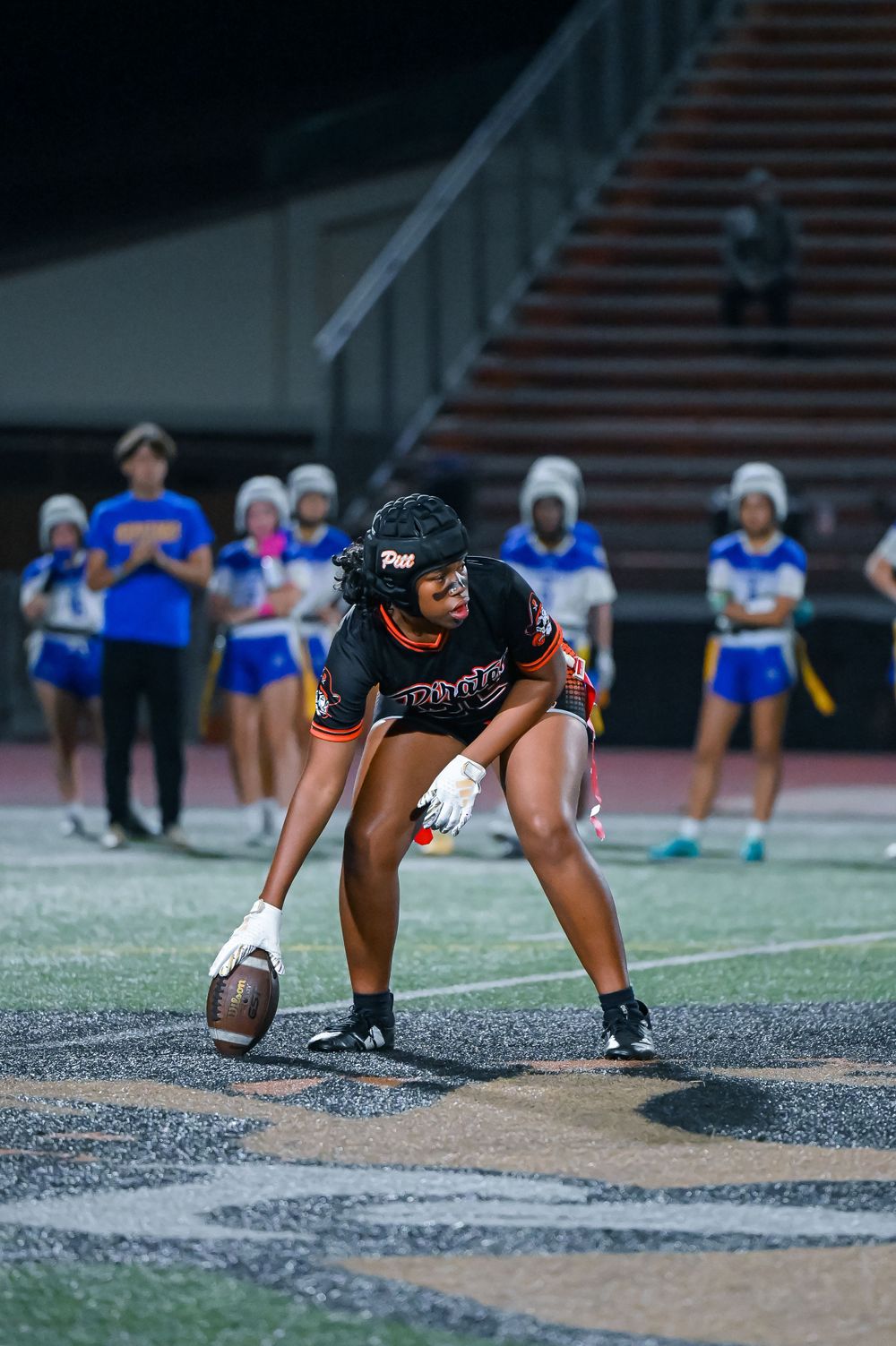 Football player in black uniform crouches in ready position on field with cheerleaders in background.