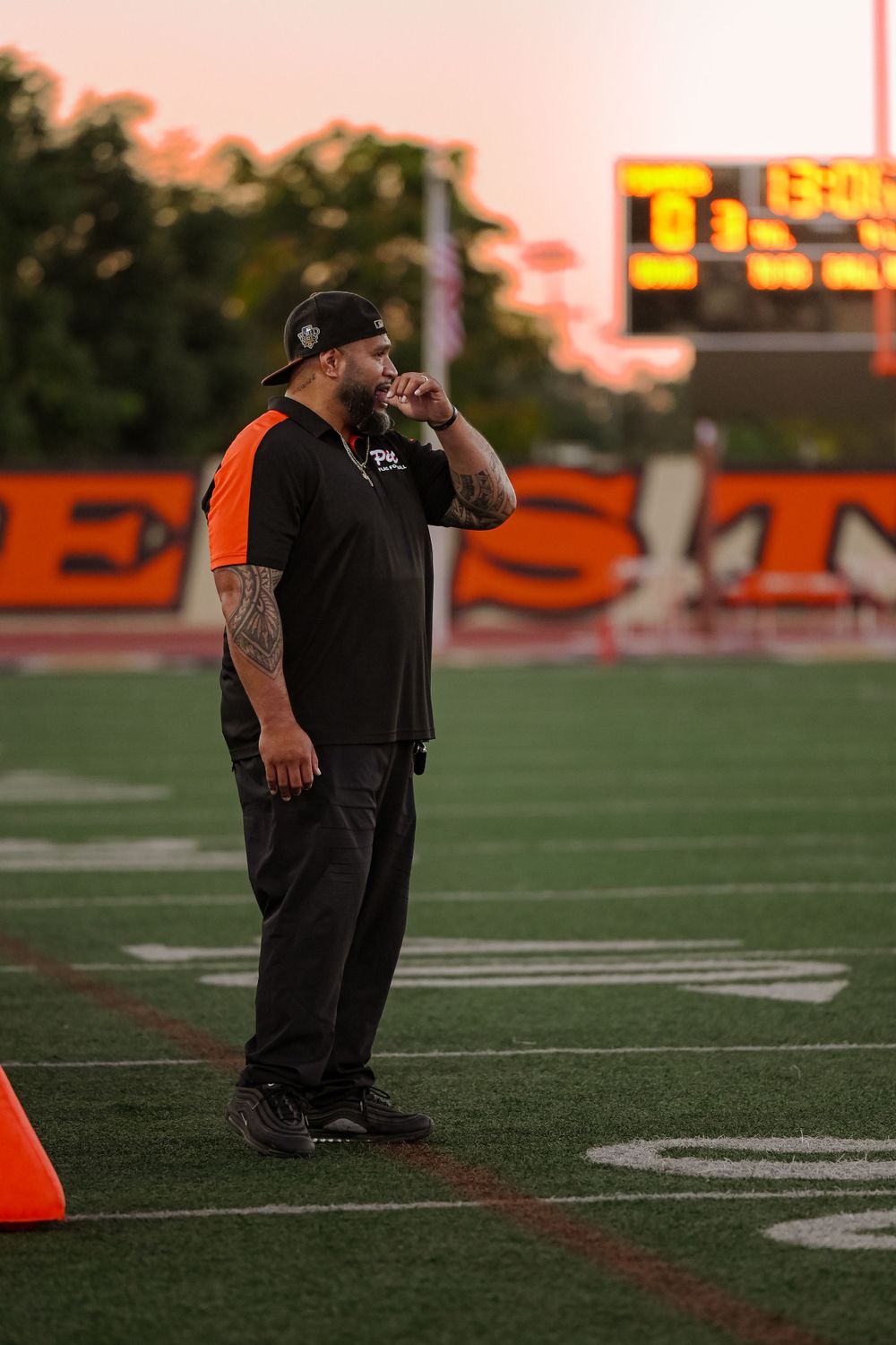 Officials in black uniforms observing from the sideline during sunset at a football game.