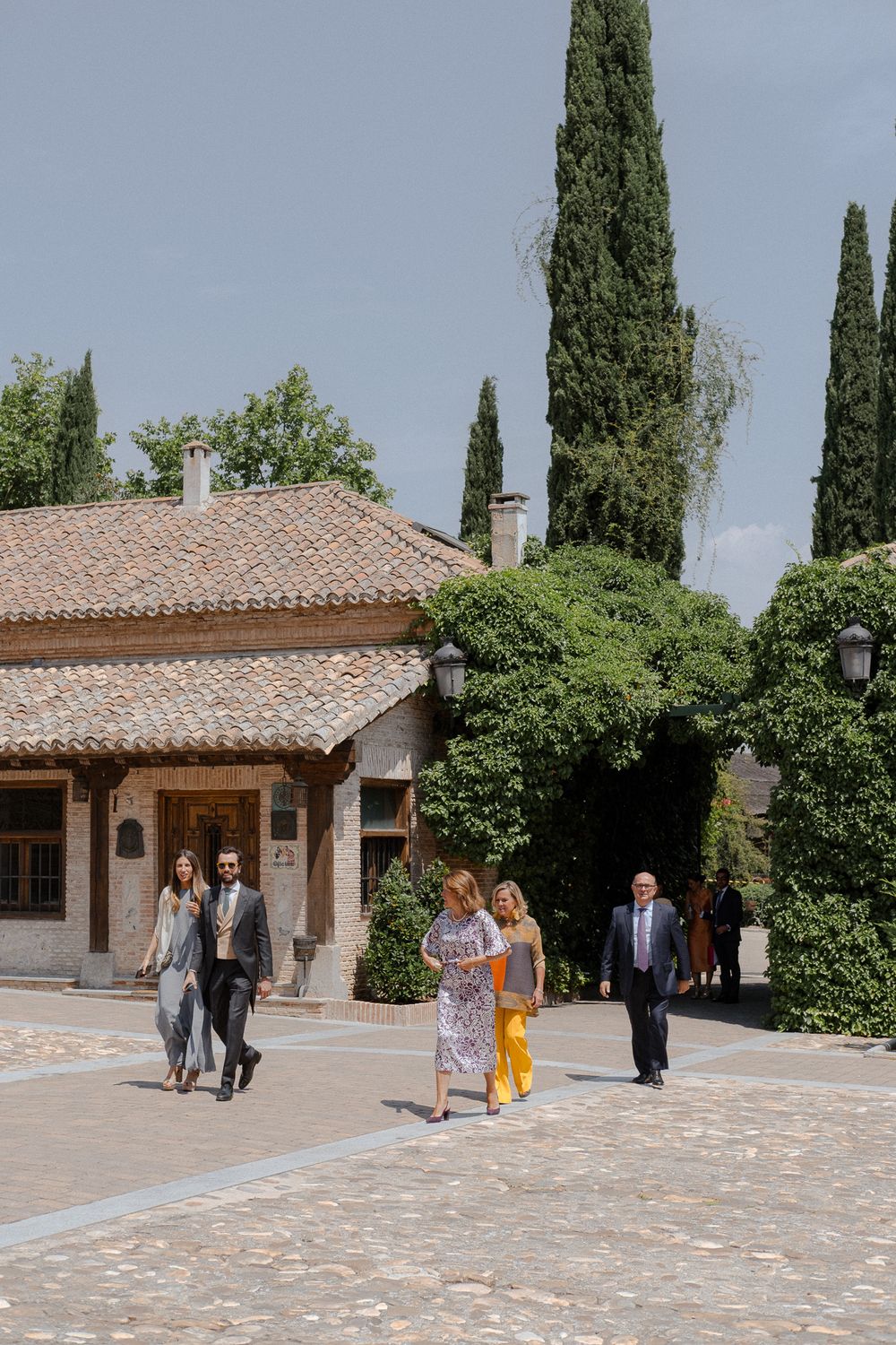 Guests walking outside a Mediterranean villa with terra cotta roof and tall cypress trees in background.