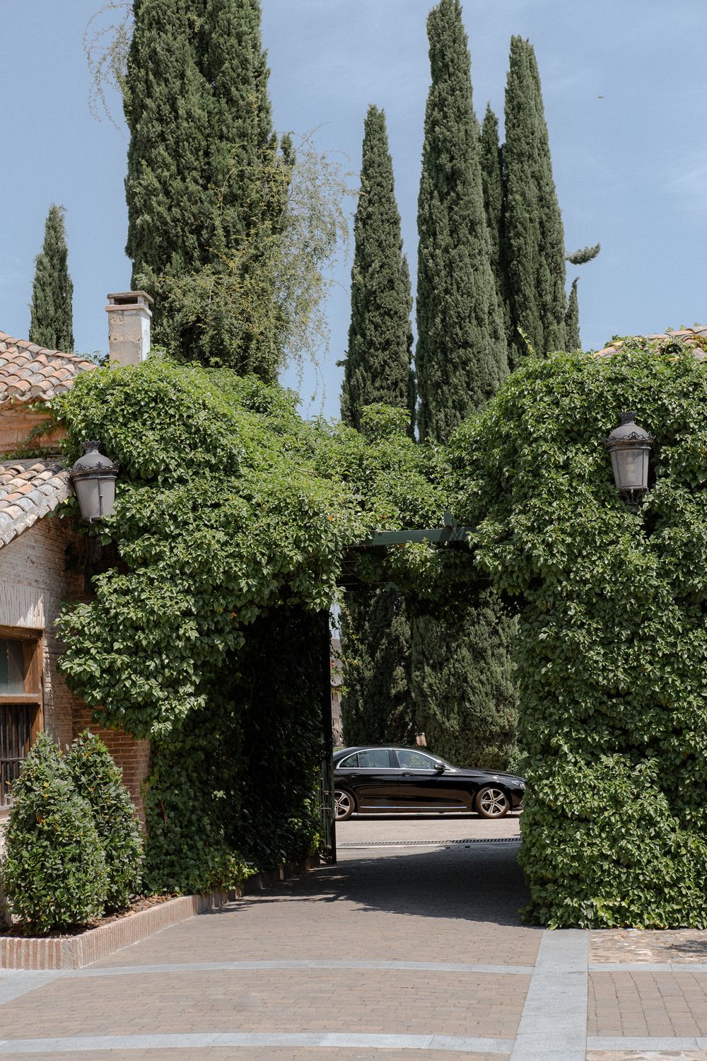 Landscaped entrance archway with tall cypress trees and parking area at Mediterranean style venue.