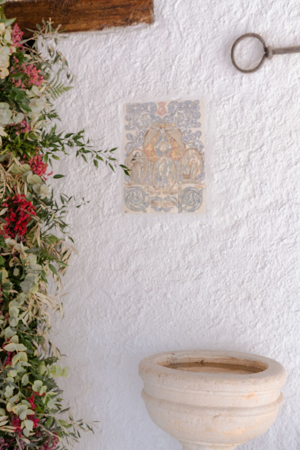 Decorative holy water font and religious artwork on white stucco wall adorned with floral arrangements.
