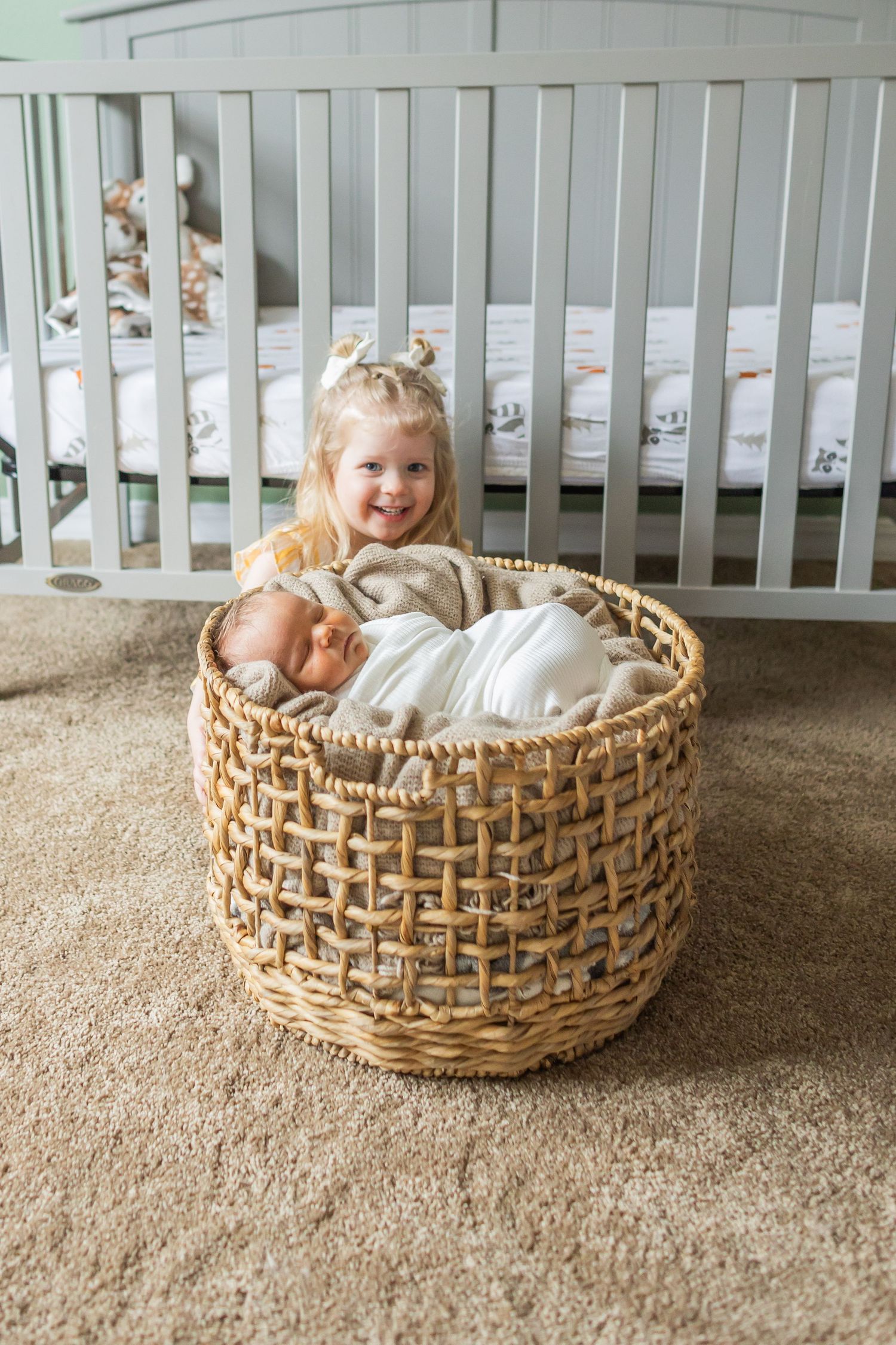 Child sits in large woven storage basket filled with blankets on carpeted floor near white crib.