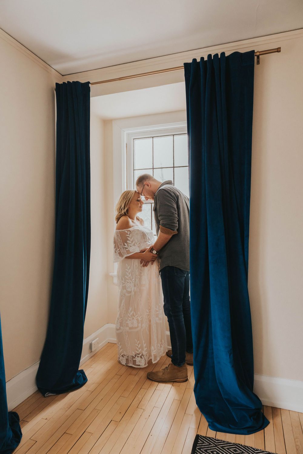 A couple shares an intimate moment by a window framed by dark blue curtains in a sunlit room with hardwood floors.