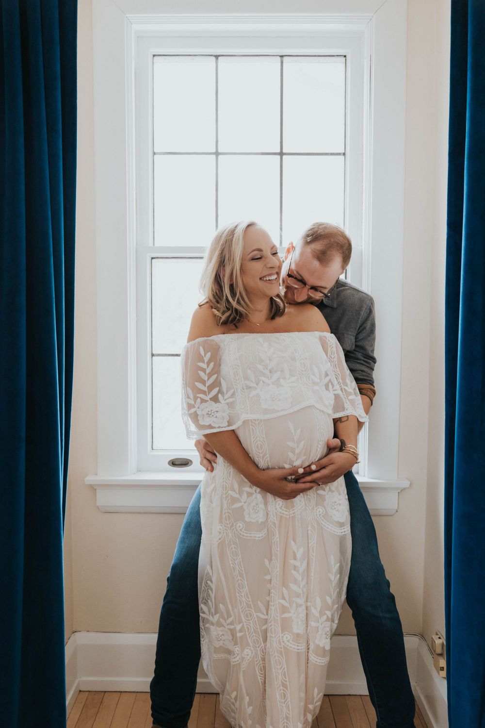 Expecting couple shares an intimate moment by a window with blue curtains as they embrace in a white lace dress.