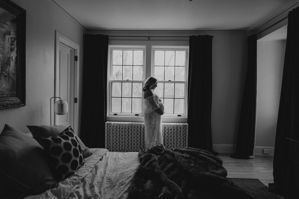 A silhouette stands by a window in a moody black and white bedroom interior with rumpled bedding in the foreground.