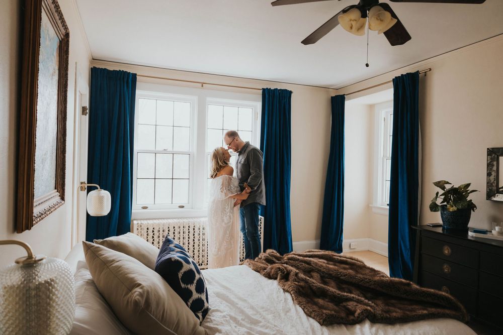 Couple standing by window in cozy bedroom with teal curtains and unmade bed.