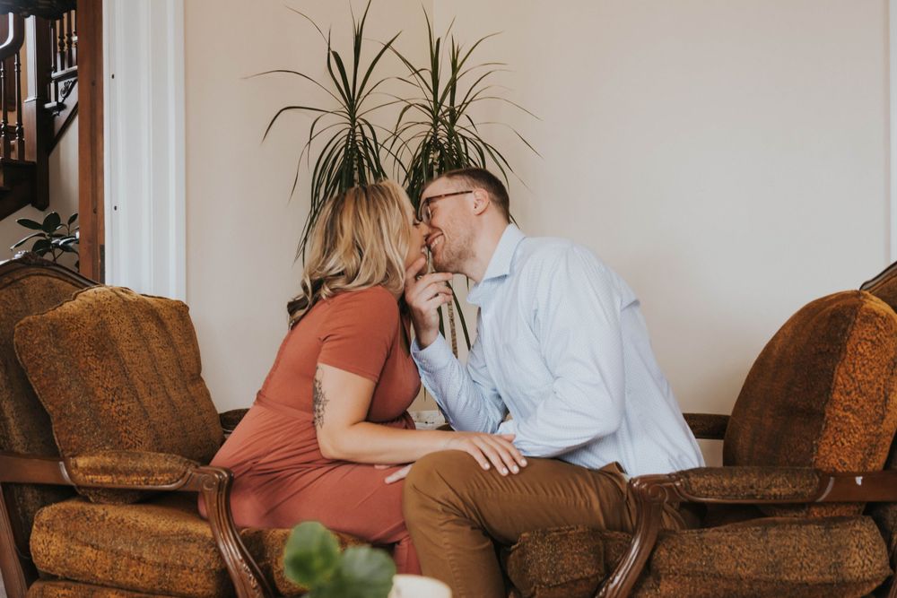 Two people share an intimate moment sitting together on a brown leather couch with potted plants in the background.
