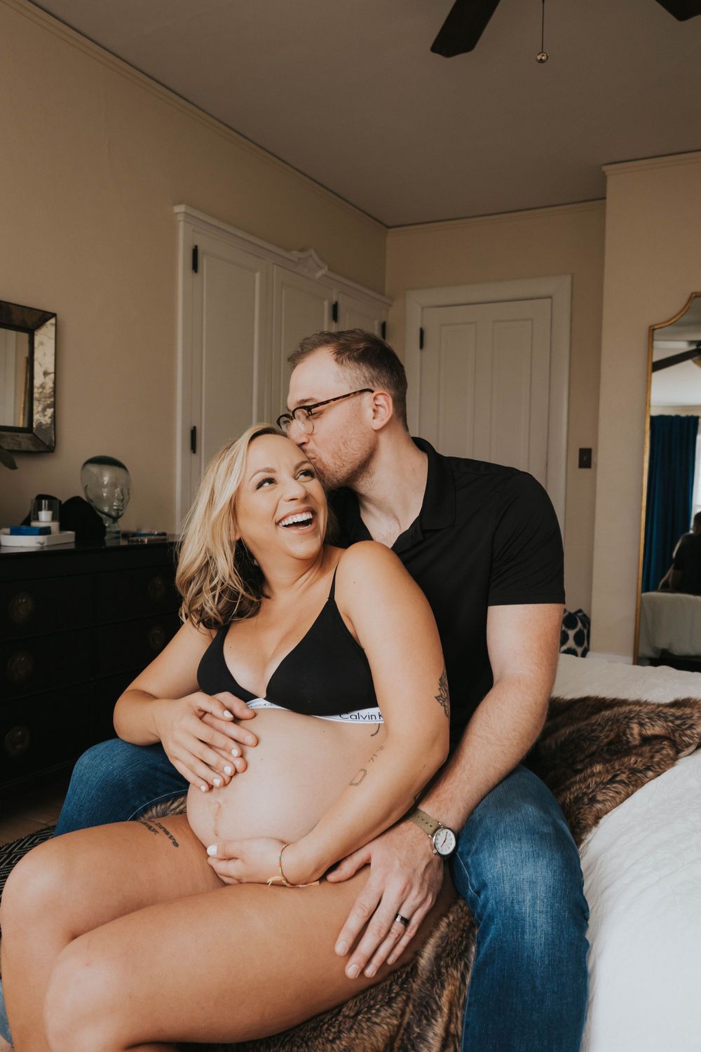 A couple shares an intimate, joyful moment together on a couch during a maternity photoshoot in warm indoor lighting.