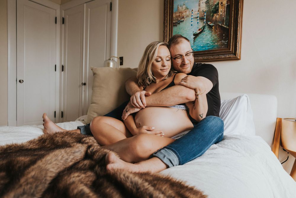 An intimate maternity photo shows a couple embracing on a bed with a brown fur blanket and artwork on the wall.