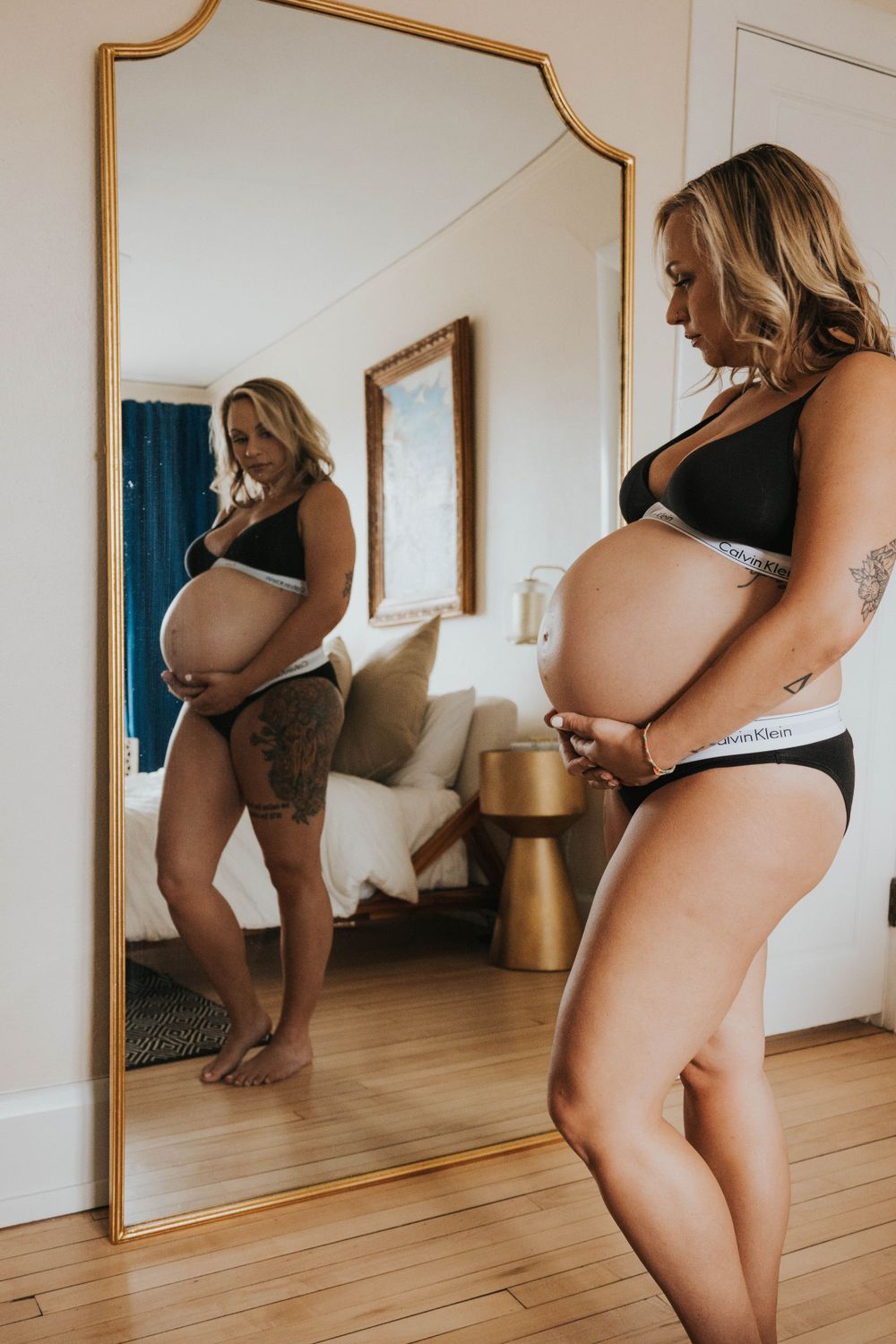 Side view of pregnant person in black underwear looking at reflection in full-length mirror in bedroom.