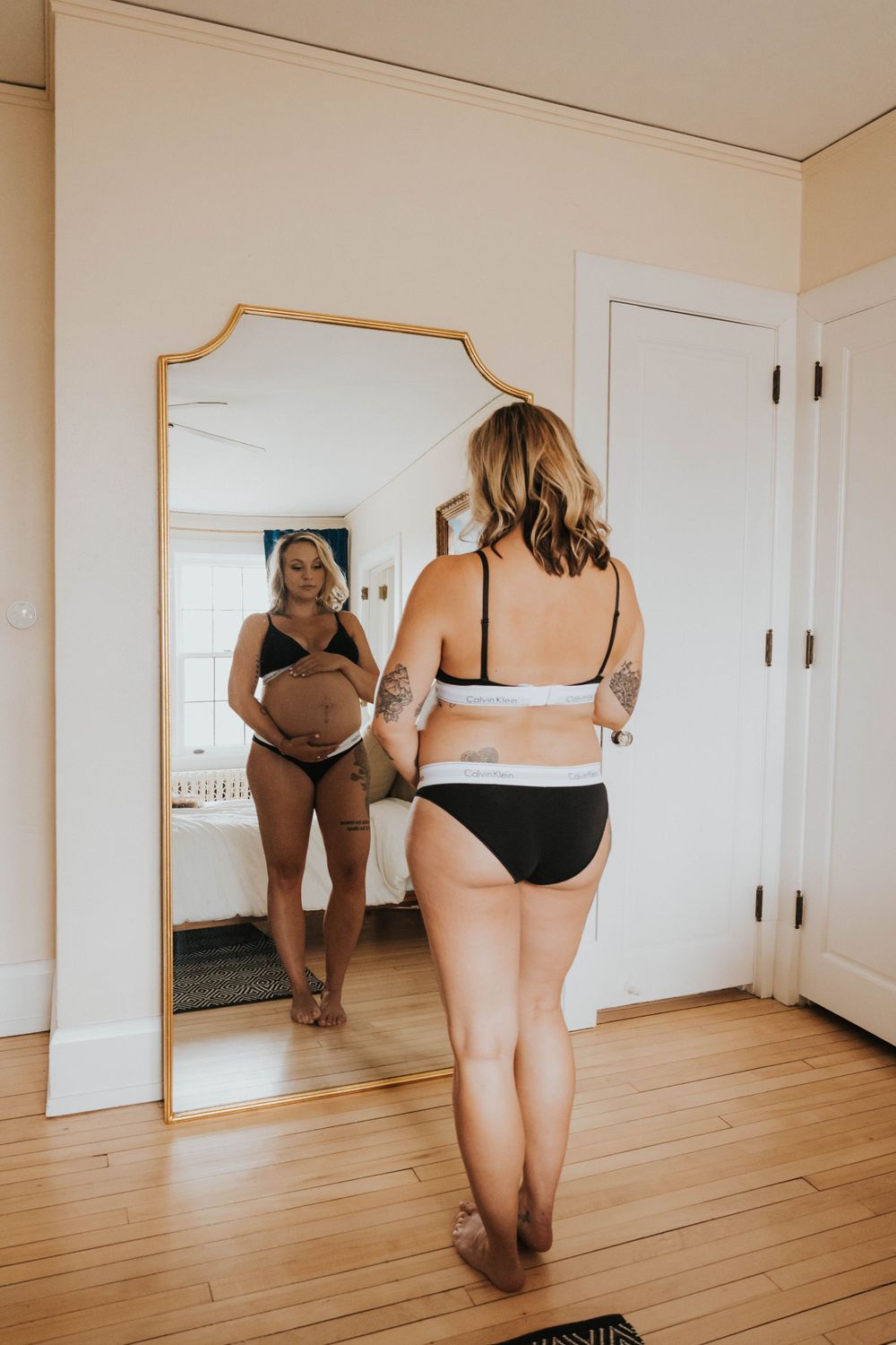 Reflection of someone wearing black underwear standing in front of a gold-framed full length mirror on hardwood floors.