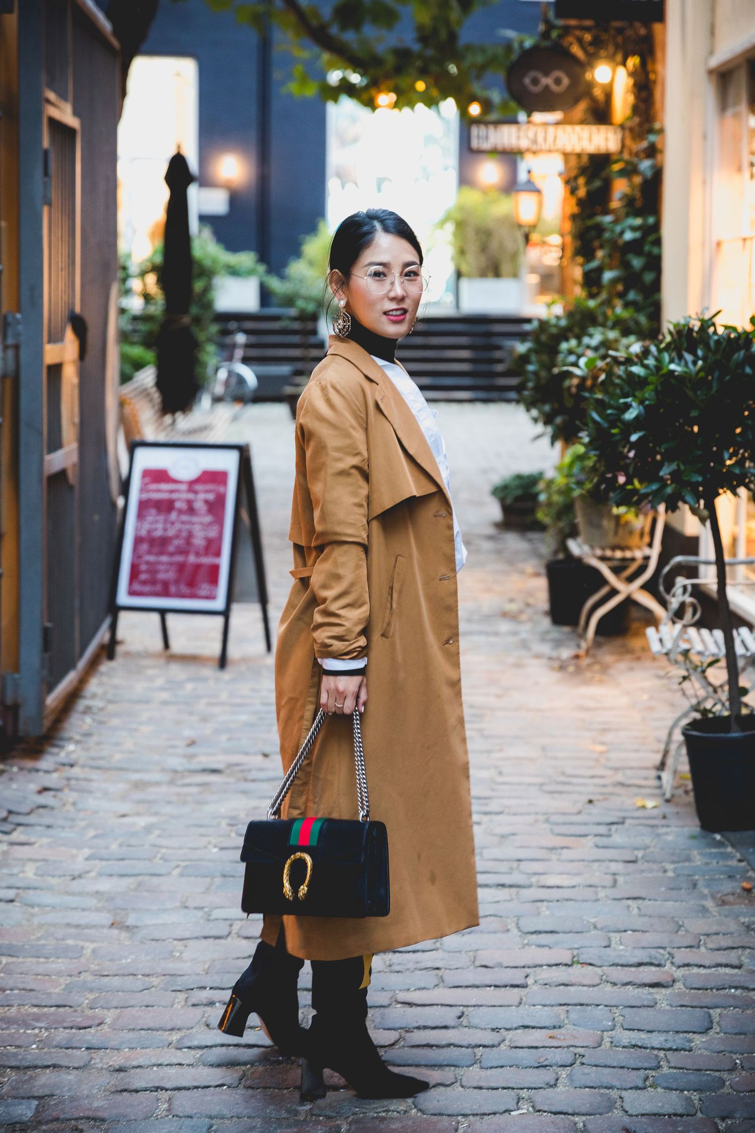 Stylish woman in tan trench coat poses with handbag on cobblestone street.