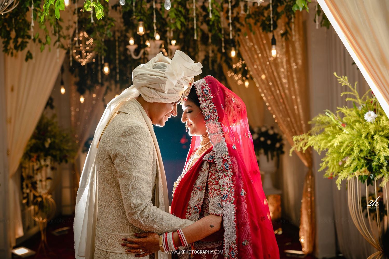 A traditional South Asian wedding ceremony under a decorated mandap with string lights and greenery overhead.
