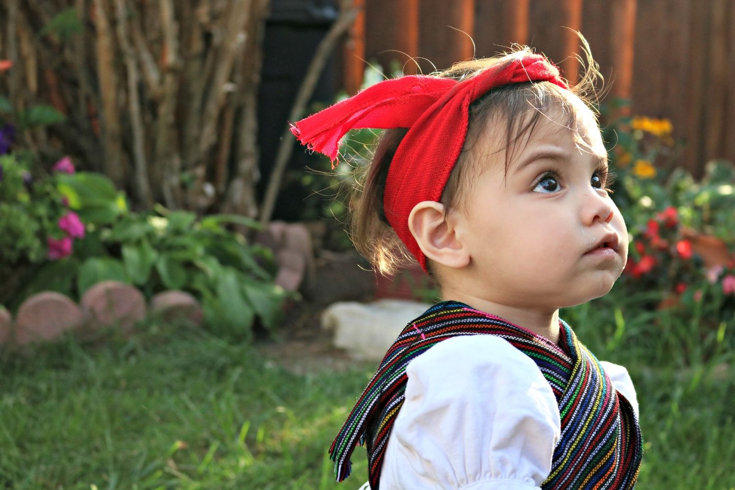 Child in white shirt and red headband looking up in a garden setting.