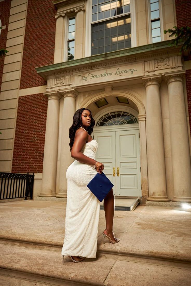Graduate in a white formal dress poses elegantly on stone steps in front of a classic brick building entrance.
