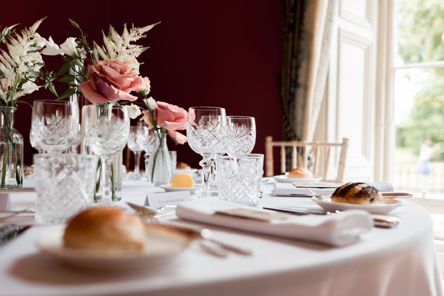 Elegant table setting with crystal glassware, white linens, and pink floral centerpiece in front of a burgundy wall.