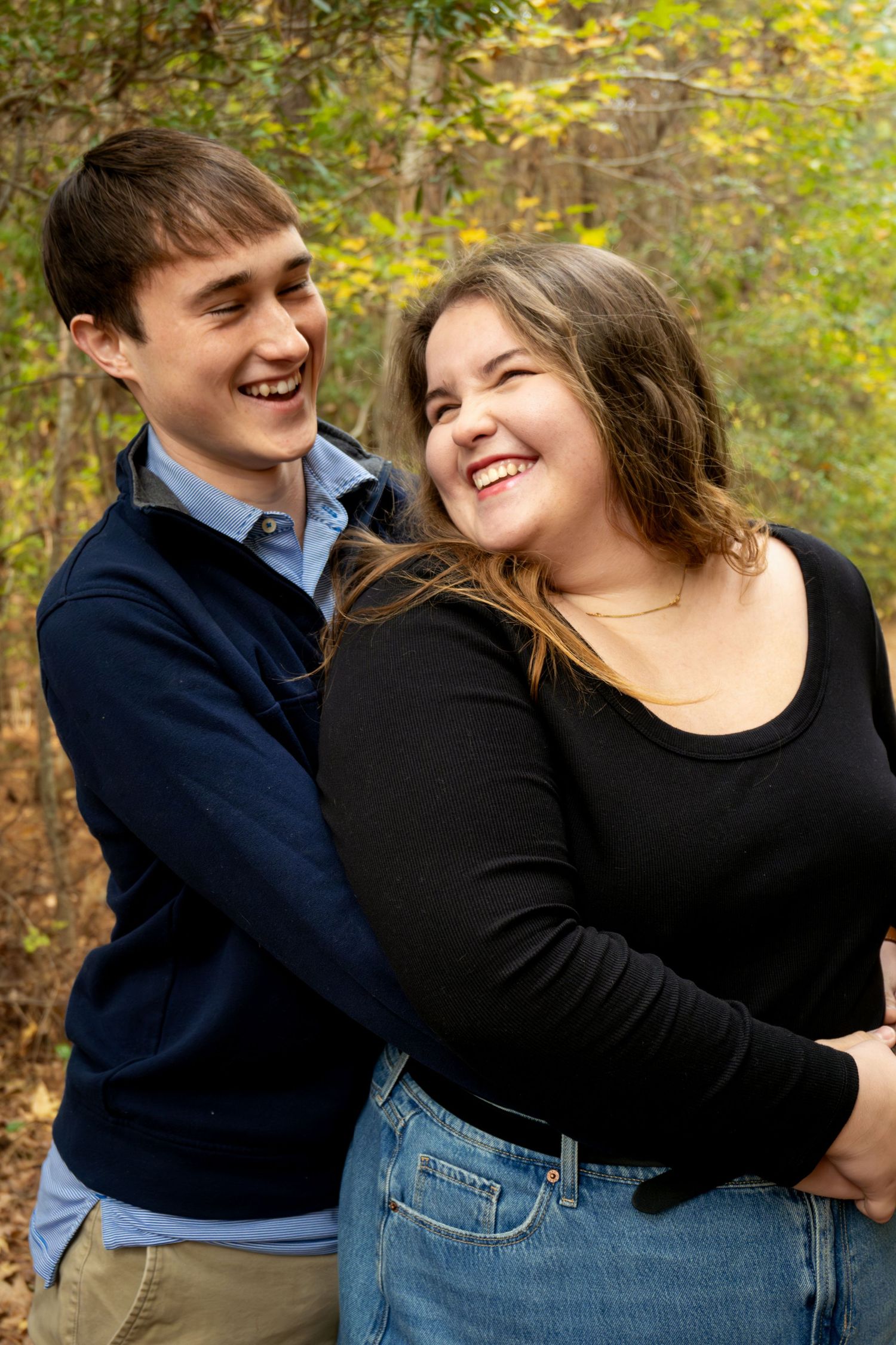 A happy couple laughs together outdoors during an autumn photoshoot in a wooded setting.