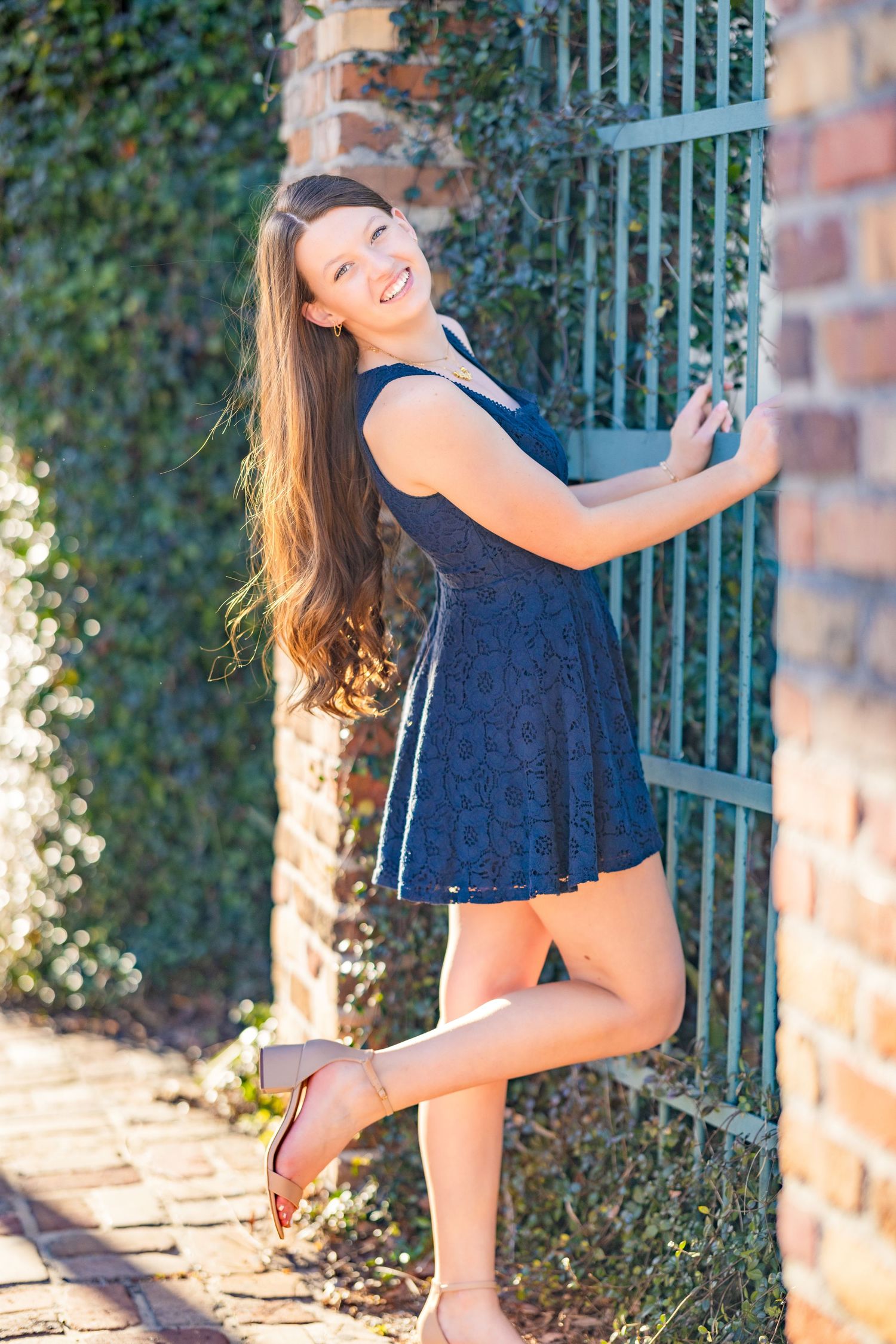 A person in a navy blue dress poses playfully against a brick wall with an iron gate and ivy in the background.