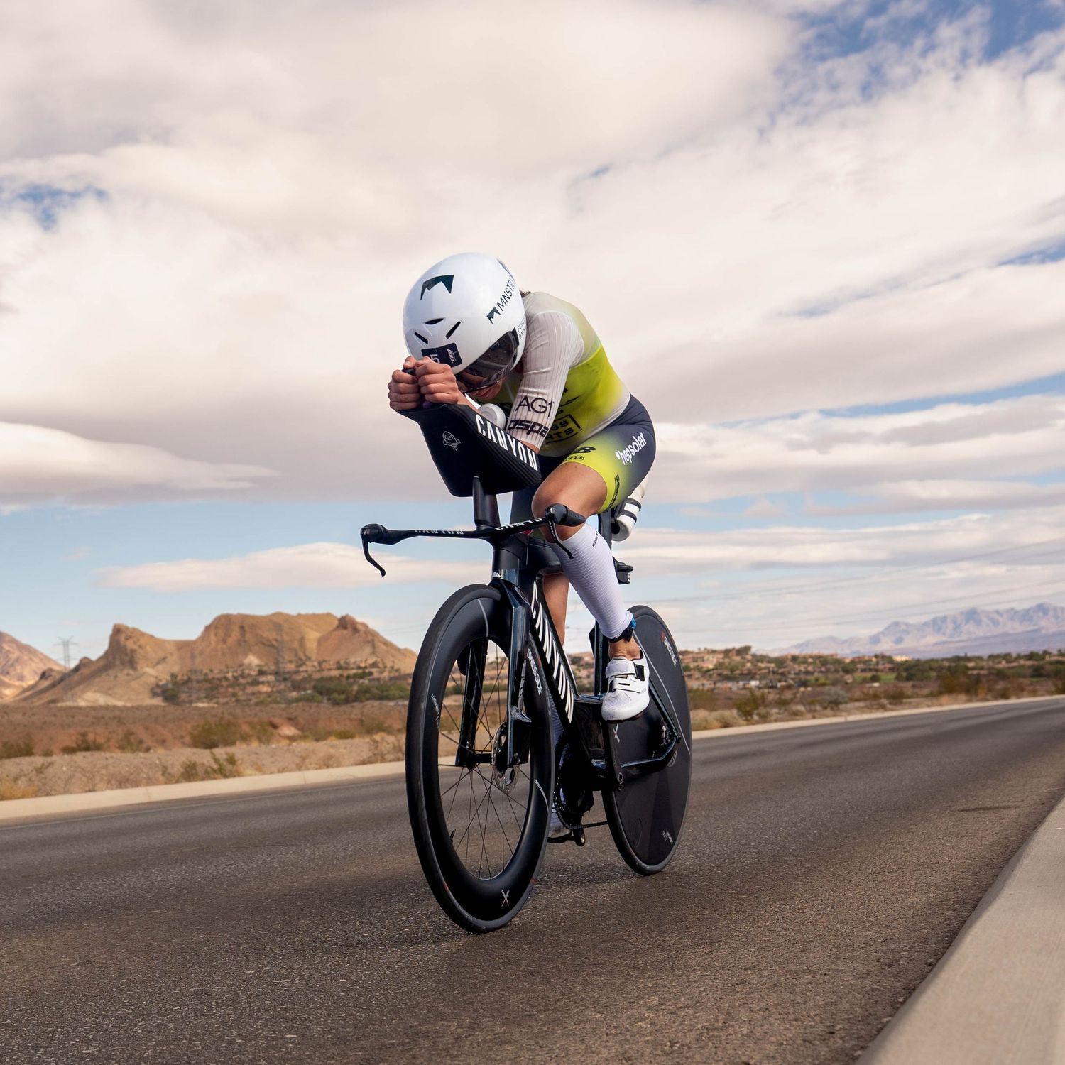 Cyclist in aerodynamic position rides time trial bike through desert landscape under cloudy skies.