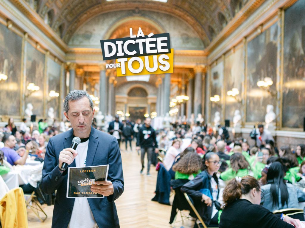 Event organizer walks through an ornate hall carrying materials during the Dictee Pour Tous spelling competition.
