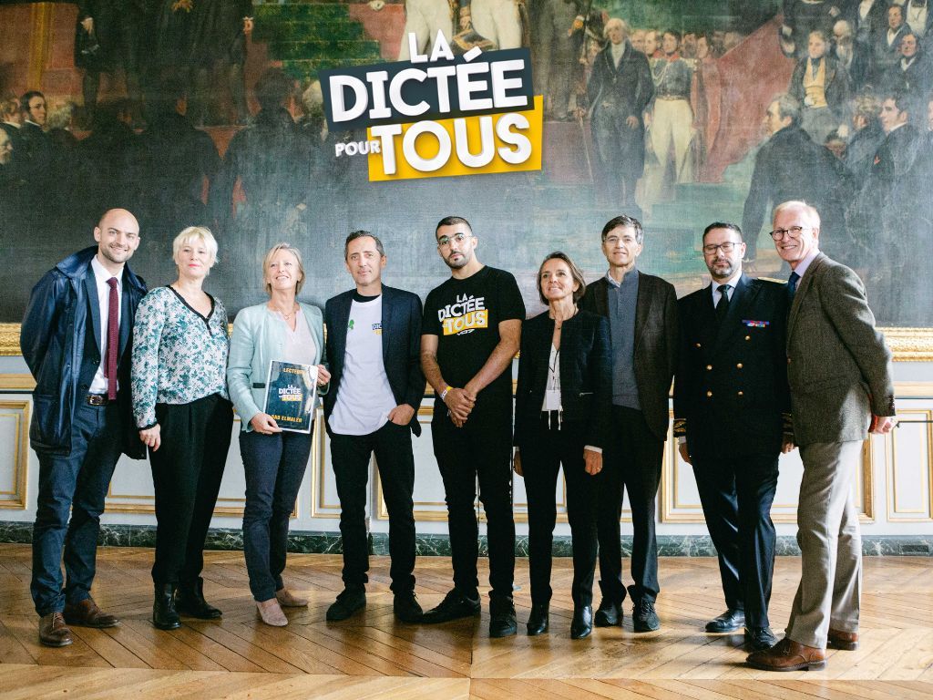 Group of people standing together indoors with the 'Dictee pour tous' logo displayed on the wall behind them.